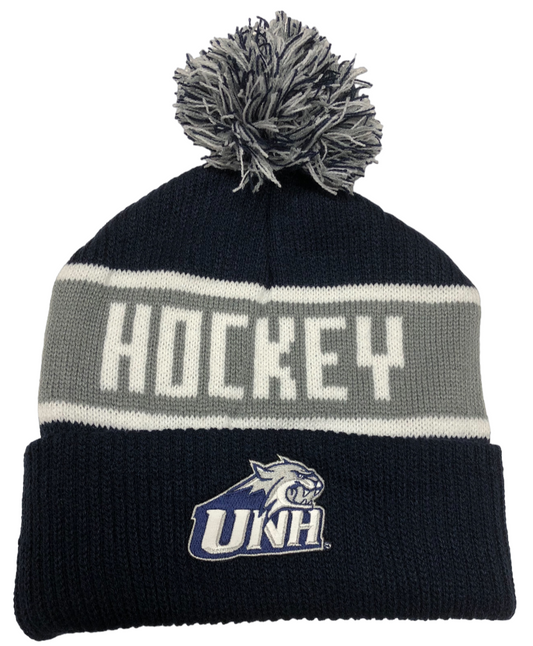 A navy knit winter hat with a white and navy fringe pom on top. The UNH Cathead logo is on the folded brim. The hat has two thin horizontal white stripes bordering a large grey horizontal stripe in the middle of it. Inside the grey stripe, the word "Hockey" is stitched in white thread in all capital letters.